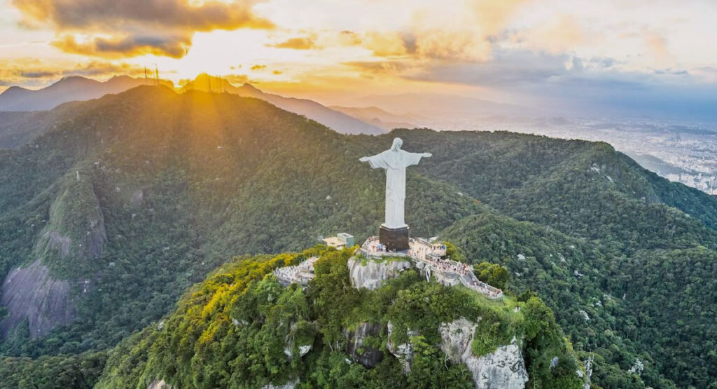 Cristo Redentor de braços abertos, foto vista do alto, ao fundo o por do sol.